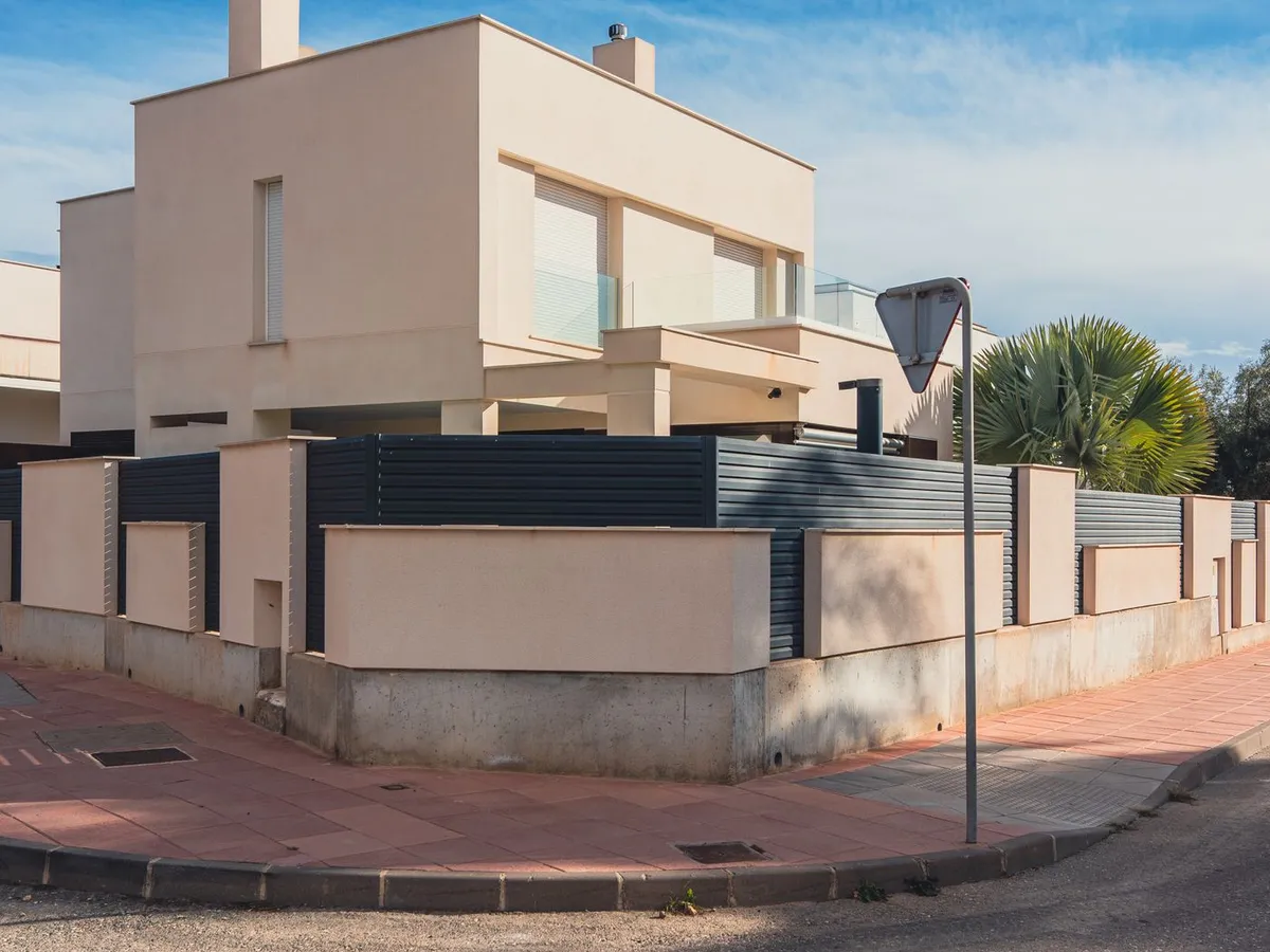 A stucco-finished masonry wall with square pillars and dark metal panels on top, wrapping a corner lot
